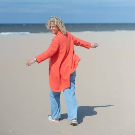 Leven na kindverlies - Desirée van Nieuwenhoven Desirée van Nieuwenhoven wandelend op het strand in een koraalrode jas